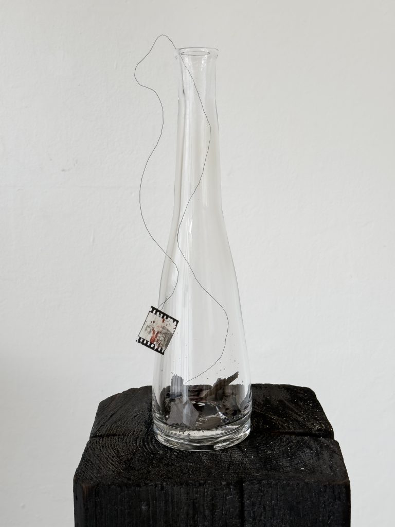 Henrik Strömberg, wishful send off 02, 2026 - handblown glass bottle, burned newspaper, metal wire and overpainted film positive 37 x 11 x 7 cm